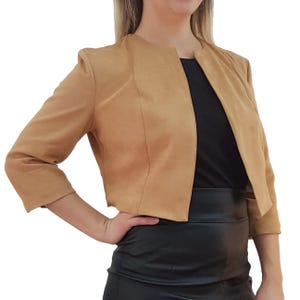 May include: A tan, cropped jacket with three-quarter sleeves. The jacket has a collarless design and a tailored fit. The fabric appears to be a suede or faux suede material. The jacket is worn over a black top and a black skirt.