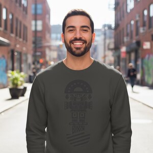May include: A dark gray sweatshirt featuring a stylized geometric design in a darker shade. The design includes a semi-circular top with star-like shapes, a central geometric figure, and a striped bottom. The person is smiling.