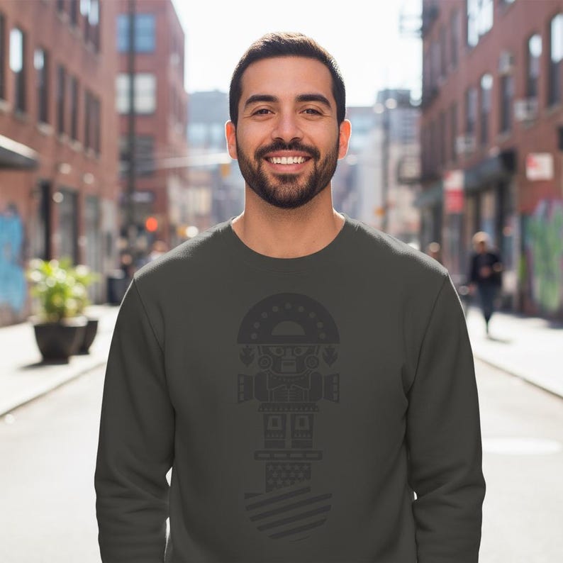 May include: A dark gray sweatshirt featuring a stylized geometric design in a darker shade. The design includes a semi-circular top with star-like shapes, a central geometric figure, and a striped bottom. The person is smiling.