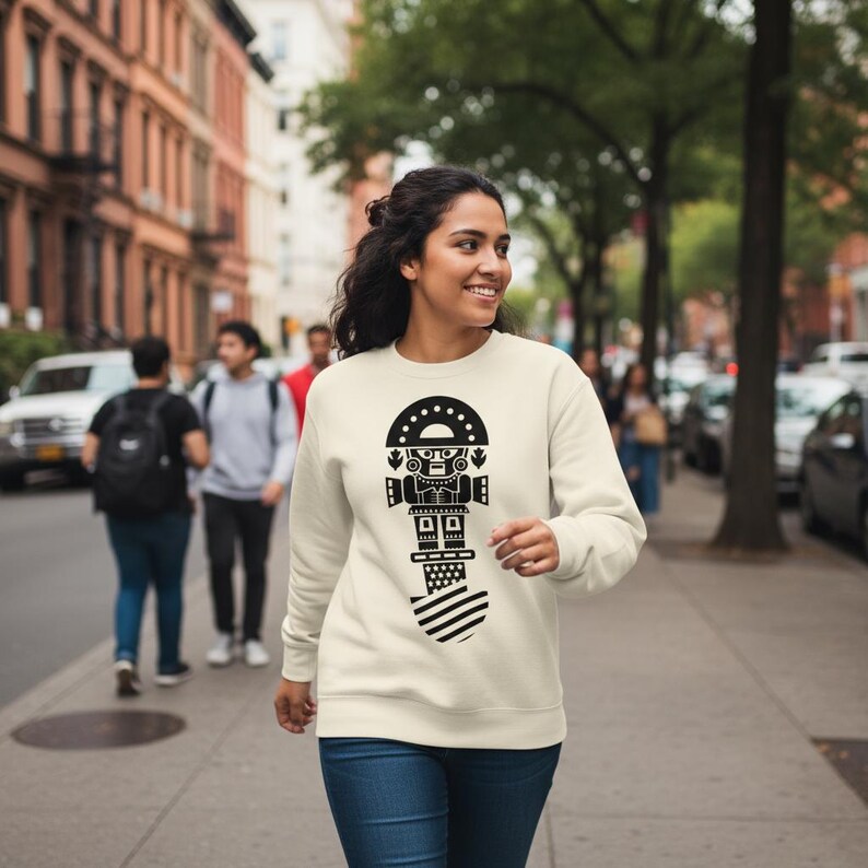 May include: Cream-colored sweatshirt with a black graphic design. The design features a stylized figure with a dotted semi-circle at the top, a rectangular body, and a striped base. The sweatshirt is being worn by a person walking on a city sidewalk.
