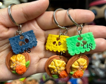 May include: Three keychains featuring a traditional Mexican sweet bread, pan dulce, with a colourful paper cutout charm. The charms are blue, yellow, and green.