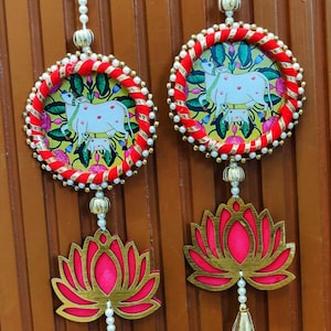 May include: Two decorative hanging ornaments. Each features a circular design with a cow illustration, surrounded by red and gold trim. Below, a pink lotus flower with gold accents hangs from a string of pearls.