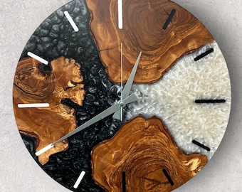May include: A round wooden clock with black and white resin accents. The clock face has a unique design with a mix of wood grain and resin. The clock hands are silver.