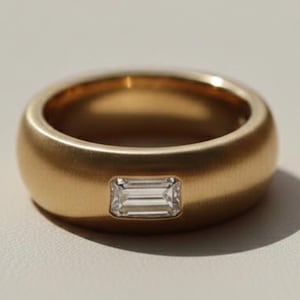 Peut inclure: Une bague en or avec une finition mate. La bague présente une pierre précieuse rectangulaire et transparente sertie au centre. L'anneau est épais et arrondi, créant une esthétique moderne et minimaliste.