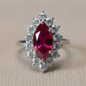 May include: A silver ring featuring a marquise-cut, deep red gemstone surrounded by a halo of smaller, round, clear gemstones. The ring's design is elegant and eye-catching, suitable for special occasions.