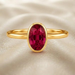 Pode incluir: Um anel de ouro com uma pedra preciosa vermelho-escura de corte oval num engaste liso. A banda é de ouro polido com um perfil arredondado, apresentando um design simples que destaca a cor vibrante da pedra central do anel.