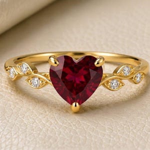 Op de afbeelding: Een gouden ring met een centrale hartvormige, donkerrode edelsteen die door klauwen wordt vastgehouden. De band is aan beide zijden ontworpen met een rankachtig motief, geaccentueerd met kleine, ronde, helderwitte stenen in de metalen blaadjes.