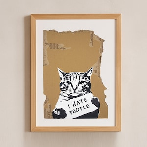 Könnte beinhalten: Gerahmter Kunstdruck mit einer schwarz-weißen Katze, die ein Schild mit der Aufschrift "I HATE PEOPLE" hält. Das Kunstwerk ist vor einem zerrissenen Kartonhintergrund platziert, in einem hellen Holzrahmen gerahmt und an einer neutralen Wand montiert.