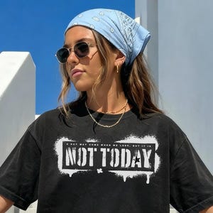 May include: Black t-shirt with the text "NOT TODAY" in a white, distressed font. The shirt has short sleeves and a crew neck. The image shows a person wearing the shirt, a light blue bandana, and sunglasses.