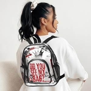 May include: Clear backpack with black trim and straps. The backpack features a graphic design with a crown, stars, and the text "DID YOU SEE MY BAG?" in red. It has mesh side pockets and a zippered front pocket.