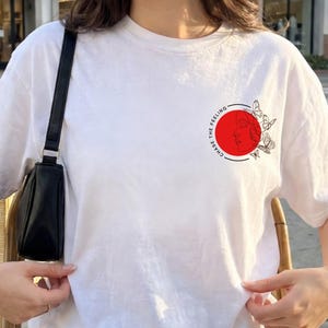K&ouml;nnte beinhalten: Wei&szlig;es T-Shirt mit einem kreisf&ouml;rmigen Aufdruck auf der Brust. Der Aufdruck zeigt einen roten Kreis mit einer schwarzen Umrandung eines Gesichts und Schmetterlingen sowie den Text "CHASE THE FEELING" in einem kreisf&ouml;rmigen Muster. Das Shirt hat kurze &Auml;rmel.