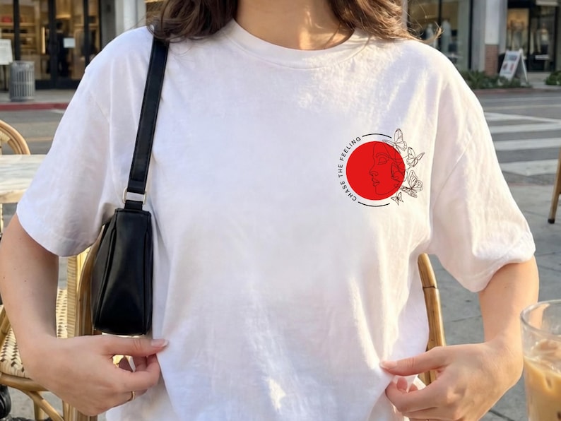 K&ouml;nnte beinhalten: Wei&szlig;es T-Shirt mit einem kreisf&ouml;rmigen Aufdruck auf der Brust. Der Aufdruck zeigt einen roten Kreis mit einer schwarzen Umrandung eines Gesichts und Schmetterlingen sowie den Text "CHASE THE FEELING" in einem kreisf&ouml;rmigen Muster. Das Shirt hat kurze &Auml;rmel.