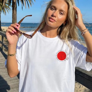 K&ouml;nnte beinhalten: Wei&szlig;es T-Shirt mit einem roten Kreis-Grafik mit Schmetterlingen und dem Text "CHASE THE FEELING". Die Person h&auml;lt eine braune Sonnenbrille. Der Hintergrund zeigt einen Holzsteg und das Meer.