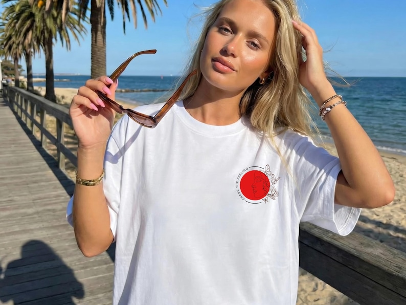 K&ouml;nnte beinhalten: Wei&szlig;es T-Shirt mit einem roten Kreis-Grafik mit Schmetterlingen und dem Text "CHASE THE FEELING". Die Person h&auml;lt eine braune Sonnenbrille. Der Hintergrund zeigt einen Holzsteg und das Meer.