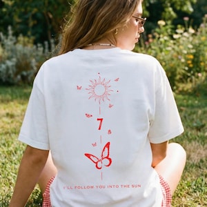 K&ouml;nnte beinhalten: Wei&szlig;es T-Shirt mit roter Sonne, Schmetterlingen und der Zahl 7 auf dem R&uuml;cken. Der Text "I'll follow you into the sun" ist unten aufgedruckt. Das T-Shirt wird im Freien getragen.