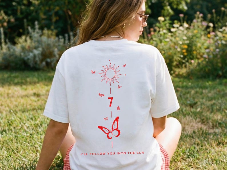 K&ouml;nnte beinhalten: Wei&szlig;es T-Shirt mit roter Sonne, Schmetterlingen und der Zahl 7 auf dem R&uuml;cken. Der Text "I'll follow you into the sun" ist unten aufgedruckt. Das T-Shirt wird im Freien getragen.