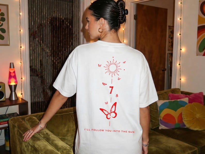 K&ouml;nnte beinhalten: Wei&szlig;es T-Shirt mit roter Grafik auf dem R&uuml;cken. Die Grafik zeigt eine Sonne, Schmetterlinge, die Zahl 7 und den Text "I'LL FOLLOW YOU INTO THE SUN". Das Shirt hat eine lockere Passform.