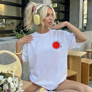 K&ouml;nnte beinhalten: Ein wei&szlig;es T-Shirt mit einem roten Kreisdesign und floralen Akzenten. Das Bild zeigt auch eine gelbe Handtasche, einen Blumenstrau&szlig; und eine Person, die gelbe Kopfh&ouml;rer und ein gelbes Armband tr&auml;gt.