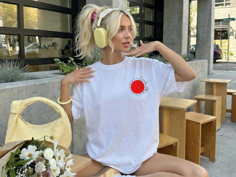 K&ouml;nnte beinhalten: Ein wei&szlig;es T-Shirt mit einem roten Kreisdesign und floralen Akzenten. Das Bild zeigt auch eine gelbe Handtasche, einen Blumenstrau&szlig; und eine Person, die gelbe Kopfh&ouml;rer und ein gelbes Armband tr&auml;gt.