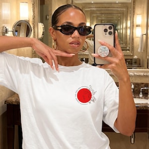K&ouml;nnte beinhalten: Wei&szlig;es T-Shirt mit einem roten Kreis-Design und dem Text "CHASE THE FEELING". Das Design beinhaltet Schmetterlinge. Die Person im Bild tr&auml;gt eine Sonnenbrille und goldene Creolen.