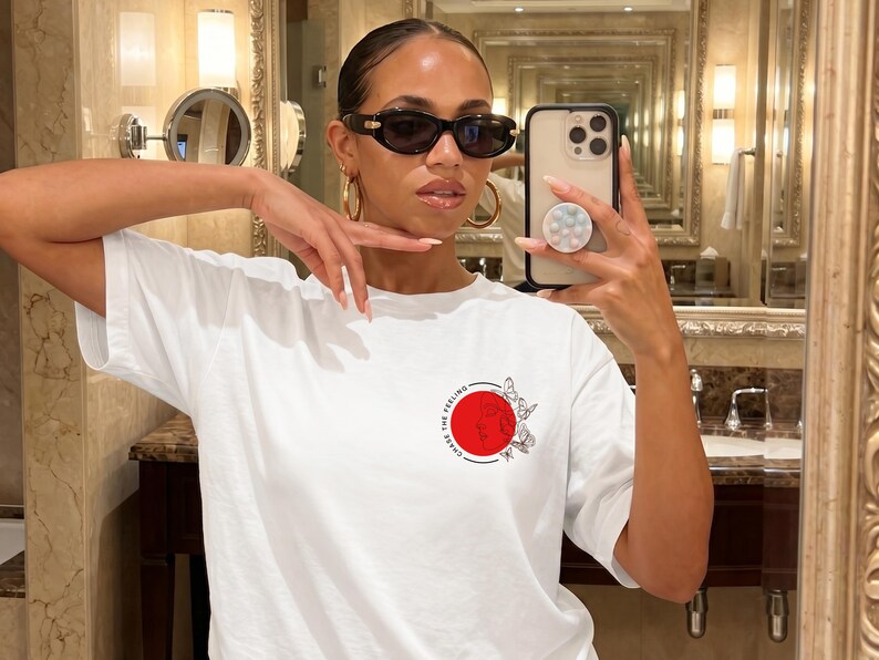 K&ouml;nnte beinhalten: Wei&szlig;es T-Shirt mit einem roten Kreis-Design und dem Text "CHASE THE FEELING". Das Design beinhaltet Schmetterlinge. Die Person im Bild tr&auml;gt eine Sonnenbrille und goldene Creolen.