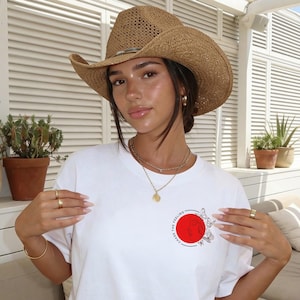 K&ouml;nnte beinhalten: Eine Person tr&auml;gt einen Cowboyhut aus Stroh und ein wei&szlig;es T-Shirt mit einem roten Kreis und einem Schmetterlingsmuster. Die Person tr&auml;gt auch Goldschmuck.