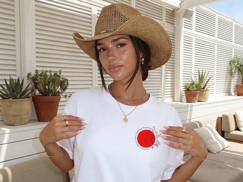 K&ouml;nnte beinhalten: Eine Person tr&auml;gt einen Cowboyhut aus Stroh und ein wei&szlig;es T-Shirt mit einem roten Kreis und einem Schmetterlingsmuster. Die Person tr&auml;gt auch Goldschmuck.