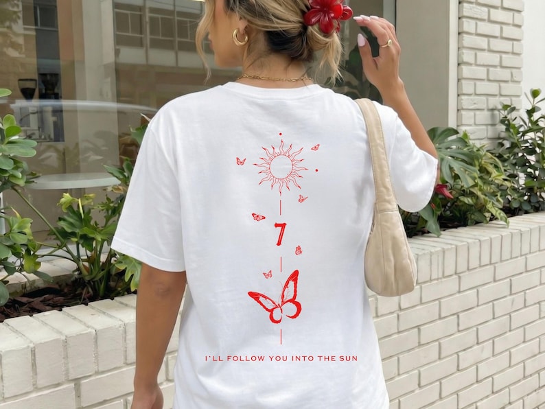 K&ouml;nnte beinhalten: Wei&szlig;es T-Shirt mit roter Grafik auf dem R&uuml;cken, die eine Sonne, Schmetterlinge, die Zahl 7 und den Schriftzug "I'LL FOLLOW YOU INTO THE SUN" zeigt. Das Shirt hat kurze &Auml;rmel und eine lockere Passform.