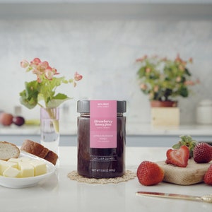 May include: A glass jar of strawberry honey jam with a pink label that reads "Strawberry Honey Jam" and "Citrus Blossom Honey". The jar is sitting on a white counter with a wooden cutting board, a plate of buttered bread, and fresh strawberries.