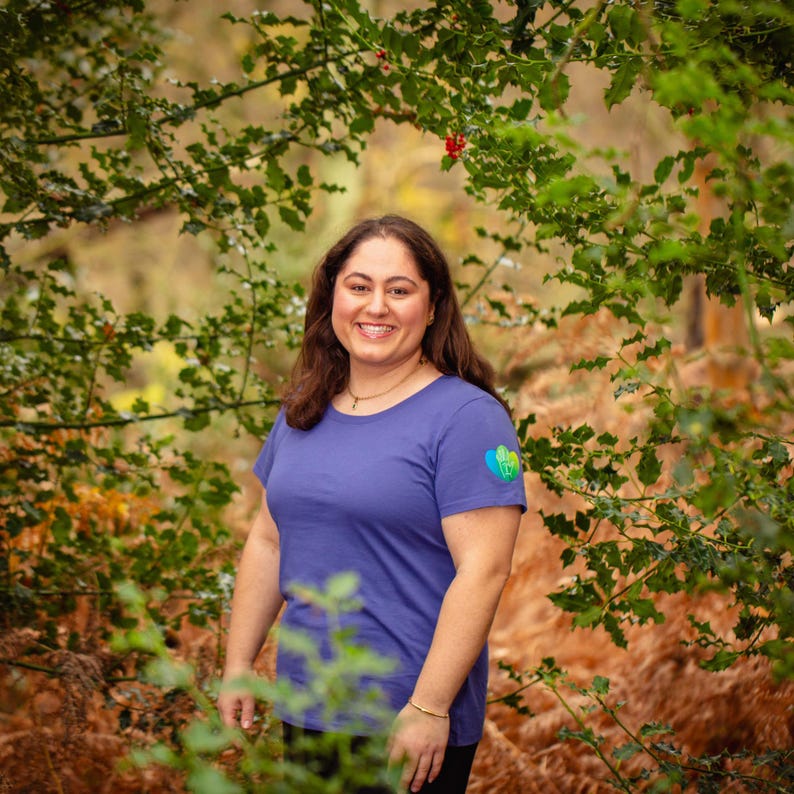 May include: A woman wearing a short-sleeved, periwinkle blue t-shirt with a small heart-shaped design on the left sleeve. The woman is smiling and standing in a forest setting, surrounded by green foliage and brown ferns.