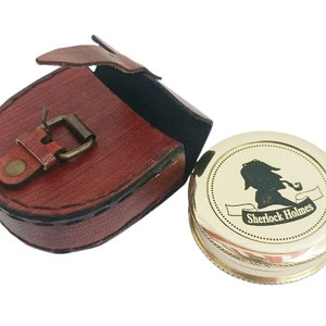 May include: A gold-colored compass with a Sherlock Holmes silhouette and text, next to a red leather case with a buckle closure. The compass has a detailed edge design.