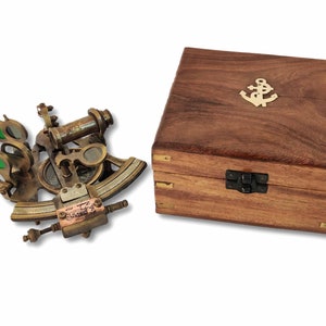 Puede incluir: Un sextante de latón antiguo con lentes verdes y una caja de madera. La caja es de madera oscura y presenta un emblema de ancla de latón en la tapa. El sextante es un instrumento de navegación.