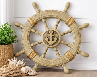 May include: A decorative ship wheel featuring a distressed light tan finish with a central circular emblem displaying a raised anchor icon. The wheel includes eight spokes and sections wrapped in natural tan rope, creating a nautical aesthetic.