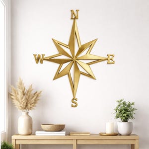 May include: A gold-colored compass rose wall decor with the cardinal directions labeled N, E, S, and W. The compass is mounted on a white wall above a wooden console table. Decorative items include a vase with dried plants, a woven bowl, and a potted plant.