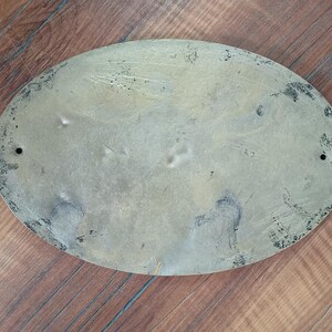 May include: An oval-shaped brass plaque with a weathered, tarnished finish. The surface displays visible scratches, dark oxidation spots, and uneven discolouration. Two small circular mounting holes are positioned on the left and right sides. captain room decor, sailor gift, sailor decor, sailor gift idea, sailing decor, sailing gift, coastal door sign, coastal door plaque, coastal wall decor, coastal home decor, coastal entry sign, coastal decor, beach house decor, seaside decor,