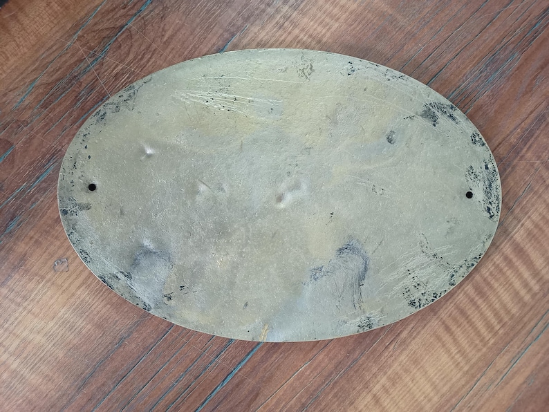 May include: An oval-shaped brass plaque with a weathered, tarnished finish. The surface displays visible scratches, dark oxidation spots, and uneven discolouration. Two small circular mounting holes are positioned on the left and right sides. captain room decor, sailor gift, sailor decor, sailor gift idea, sailing decor, sailing gift, coastal door sign, coastal door plaque, coastal wall decor, coastal home decor, coastal entry sign, coastal decor, beach house decor, seaside decor,