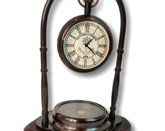 May include: A vintage clock and compass set. The clock face displays Roman numerals and the text "Victoria London 1876". The clock is suspended within a dark metal arch and sits on a dark wooden base. The compass is at the base.