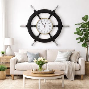 Puede incluir: Un gran reloj con forma de timón negro con detalles plateados cuelga de una pared blanca. La esfera del reloj es blanca con números romanos negros y ribete dorado. Un artículo de decoración náutica.