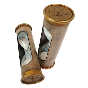 May include: Two vintage hourglasses with brass detailing and tan leather casing. The glass vials contain white sand. The hourglasses are cylindrical and have a classic design. The top and bottom of the hourglasses are brass with engraved details.