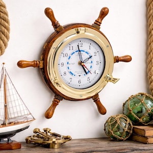 Puede incluir: Un reloj de pared de temática náutica con forma de timón de barco. El reloj tiene una esfera blanca con números negros, un bisel de latón y radios de madera. Decoración náutica.