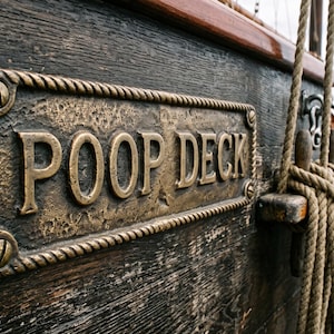 Könnte beinhalten: Eine rechteckige Bronzetafel mit seilartigem Rand und dem erhabenen Schriftzug POOP DECK, montiert an einer dunklen, verwitterten Schiffswand aus Holz. Die Tafel ist mit vier sichtbaren Schrauben befestigt und befindet sich neben dickem Tauwerk.