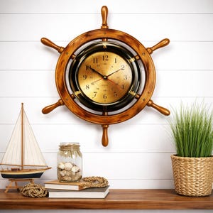 Puede incluir: Un reloj de rueda de barco de madera con esfera dorada y bisel negro. El reloj tiene un tema náutico, con un modelo de velero, un tarro de conchas y una cesta tejida con vegetación.