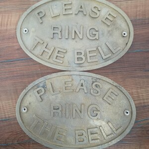 May include: Two oval-shaped brass signs with a weathered finish. Each sign features raised lettering that reads "PLEASE RING THE BELL" in a clear, sans-serif font. There are two small circular mounting holes on the left and right sides of each sign. coastal home decor, coastal entry sign, coastal decor, beach house decor, seaside decor, ocean decor, sea decor, ship decor, ship door sign, ship door plaque, ship cabin decor, maritime gift, nautical gift, nautical gift idea,