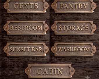 Op de afbeelding: Een verzameling van zeven rechthoekige, bronskleurige metalen bordjes met afgeronde uiteinden en schroefgaten aan de zijkanten. De bordjes zijn voorzien van verhoogde letters met de teksten GENTS, PANTRY, RESTROOM, STORAGE, SUNSET BAR, WASHROOM en CABIN.