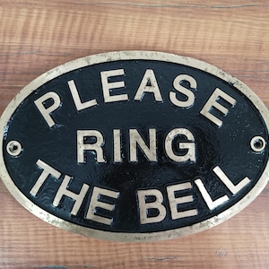 May include: An oval-shaped metal sign with a black background and a brass-coloured border. The sign features raised, metallic lettering that reads "PLEASE RING THE BELL" in three lines. There are two circular mounting holes on the left and right sides. boat cabin plaque, boat decor, boat interior decor, boat entrance sign, boat owner gift, boat gift idea, yacht door sign, yacht door plaque, yacht entry sign, yacht entry plaque, yacht wall plaque, yacht wall decor, yacht cabin sign,