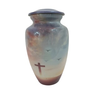 May include: A decorative urn with a sky-themed design. The urn features a cross and birds in flight against a backdrop of a sunset. The urn is a muted purple and blue color.