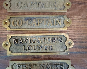 Op de afbeelding: Vier rechthoekige koperen platen met verhoogde letters en schroefgaten aan beide uiteinden. De platen zijn voorzien van de tekst CAPTAIN, CO-CAPTAIN, NAVIGATOR'S LOUNGE en FIRST MATE'S QUARTERS. Elke plaat heeft een textuurafwerking.