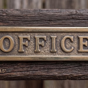 May include: A rectangular brass office sign with raised lettering reading OFFICE. The sign features a textured background, rounded ends with screw holes, and measures 5.5 inches by 1.5 inches as indicated by the text below the plaque.