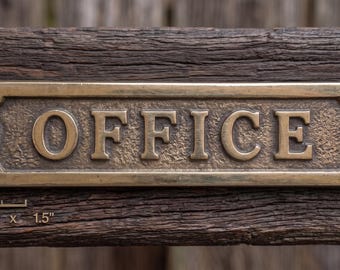 Op de afbeelding: Een rechthoekig kantoorbord van messing met verhoogde letters die OFFICE spellen. Het bord heeft een getextureerde achtergrond, afgeronde uiteinden met schroefgaten en meet 14 cm bij 3,8 cm zoals aangegeven door de tekst onder het bord.