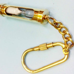 May include: A gold-colored hourglass keychain. The hourglass is encased in a cylindrical gold metal frame with a chain and clasp. The hourglass contains white sand and a clear glass chamber.